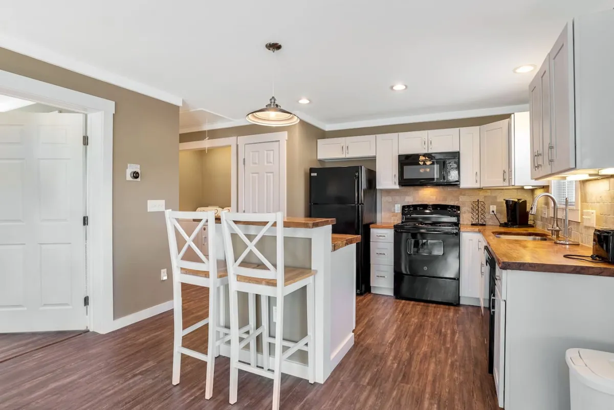 This image showcases a spacious kitchen with a central island and bar stools, flanked by sleek black appliances and surrounded by light-colored cabinets. The setup invites both culinary creativity and social interaction, with enough room for multiple activities to take place—be it cooking, chatting, or casual dining.