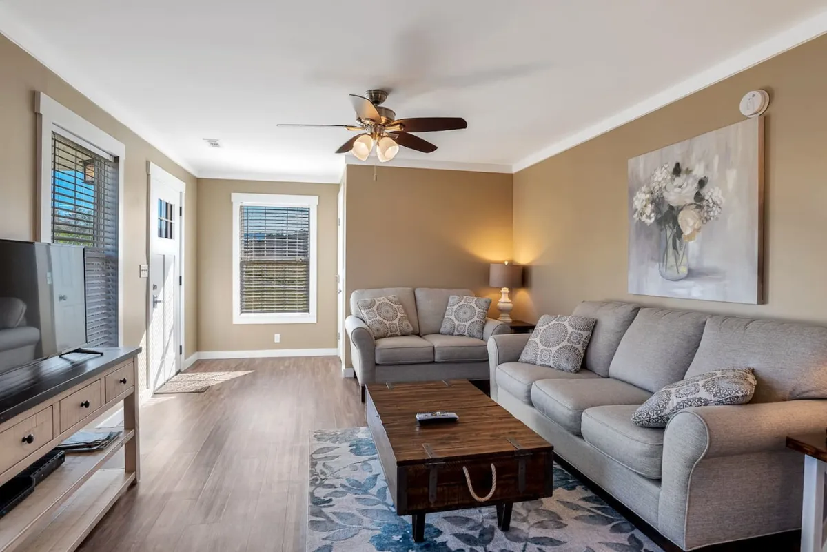 A sunlit living room with a plush sofa, inviting coffee table, large window letting in natural light, stylish artwork on the walls, and a smart TV ready for movie nights. The space exudes comfort, encouraging friendly conversation or relaxing with a book.