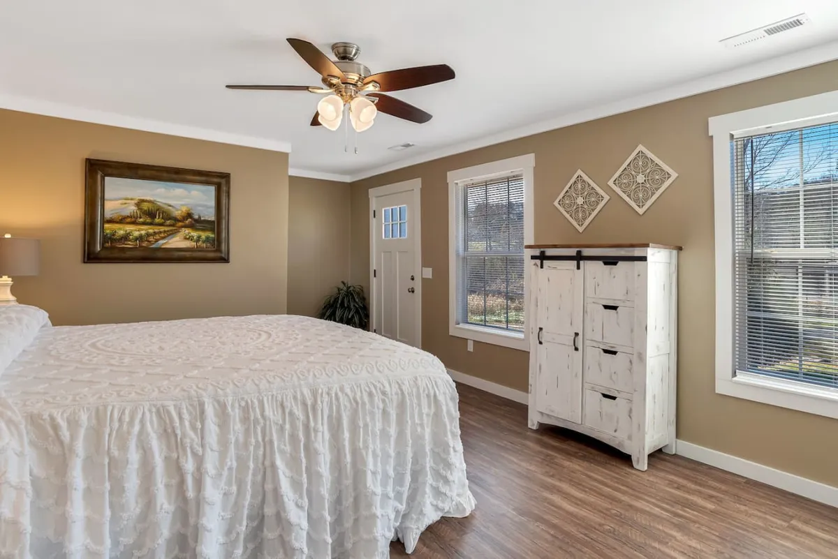 A calm, cozy bedroom featuring a bed with a white bedspread, soft blankets, wall art, a small white furniture piece, a door with a window, and a plant. Gentle natural light fills the space, creating an inviting and serene atmosphere.