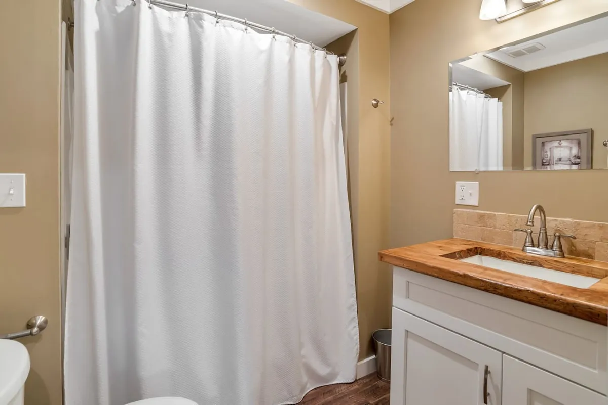 This image showcases a serene bathroom scene featuring a shower curtain, a sink set atop a wooden countertop, and a well-lit mirror. The space exudes a calming, spa-like atmosphere, emphasizing relaxation and self-care rituals.